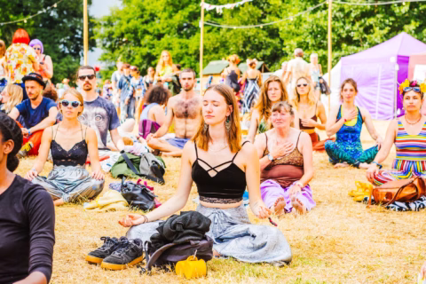 A group of people sit cross-legged on grass at an outdoor festival, meditating with eyes closed. Tents, trees, and festival-goers are in the background. The scene is colourful and relaxed.