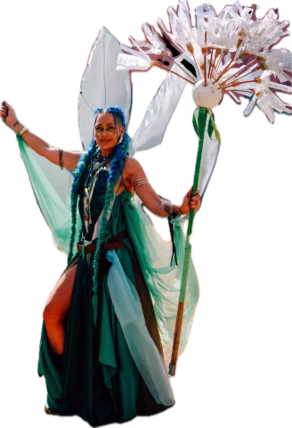 A person in a flowing green and blue costume with large leaf-shaped wings and blue hair holds a staff topped with large, white flower petals, smiling and posing with arms outstretched.