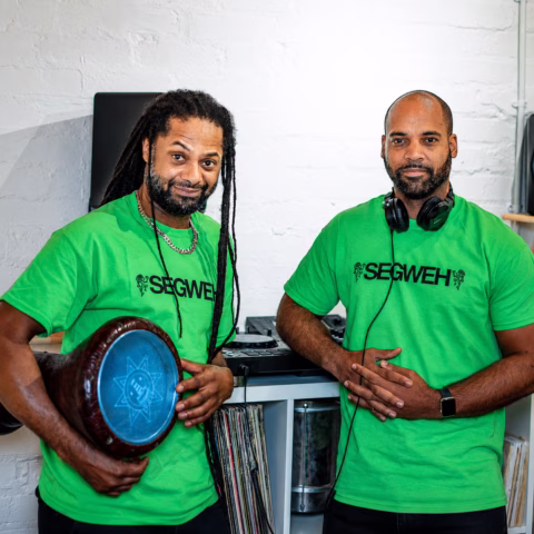 Two men wearing bright green SEGWEH shirts stand indoors. One holds a drum, the other has headphones round his neck. Vinyl records and DJ equipment are visible in the background. Both are facing the camera and smiling.