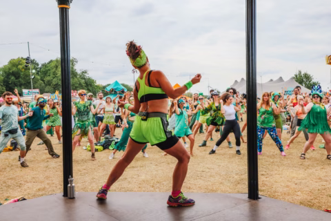 A woman in a bright green outfit leads a large group dance outdoors; participants wearing colourful clothes mimic her moves on a grassy field at what appears to be a festival.