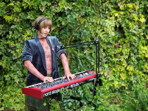 A person wearing headphones and a velvet jacket plays a red Nord Stage keyboard and sings into a microphone, performing outdoors in front of a lush, green ivy-covered wall.
