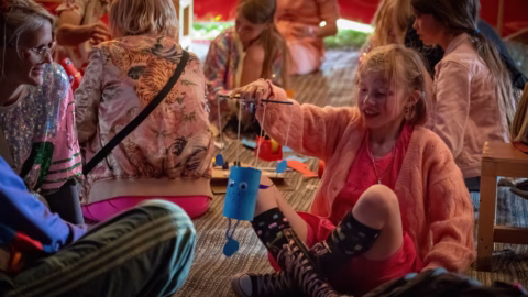 A smiling girl sits on the floor holding up a homemade blue craft with string, surrounded by other children and adults engaged in crafting activities inside a colourful, cosy setting.