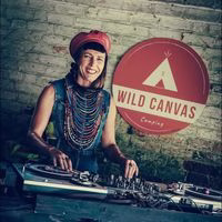 A smiling person wearing a red beret and colourful beaded necklaces stands at a DJ turntable. Behind them is a brick wall and a round red Wild Canvas sign with a white tent logo.