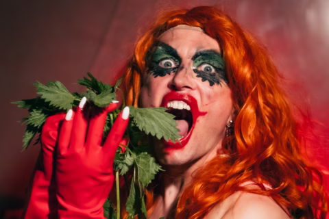 Person with long, bright red hair and dramatic green and black eye make-up, wearing red gloves, poses with mouth open near green leaves, appearing to eat them. Background is warmly lit and out of focus.