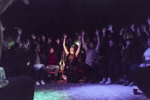 A group of people sitting in a circle with their arms raised, smiling and cheering, under colourful stage lighting in a dark room. A person in the centre appears to be leading the activity.