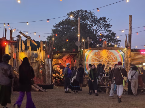 People gather at an outdoor festival in the evening, surrounded by colourful lights, wooden structures, and vibrant art. Some stand and talk, while others sit, creating a lively, communal atmosphere under fairy lights.