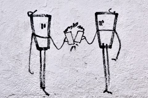 Simple black line graffiti on a white wall depicting two abstract human-like figures clinking cups together, with a small burst above the cups suggesting celebration or a toast.
