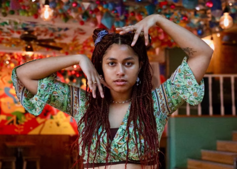 A woman with long plaited hair poses indoors, holding her hands around her face. She wears a patterned green top and stands in a colourful, festive space with hanging decorations and warm lighting.