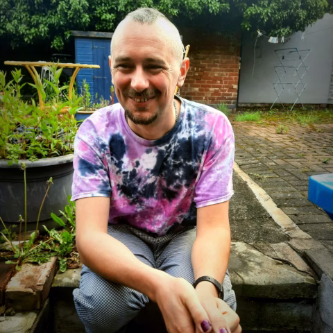 A smiling person with a shaven hairstyle and a tie-dye shirt sits on a garden step. There are plants, a blue shed, and a clothes airer in the sunny brick-paved back garden.