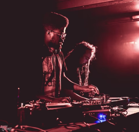 Two DJs perform at turntables in a dimly lit, red-hued room. One DJ wears glasses and headphones, while the other has long hair. Both are focused on their equipment, creating a dynamic musical atmosphere.