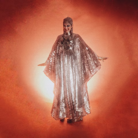 A person stands smiling with arms slightly outstretched, wearing a shimmering, metallic, full-length cape and headpiece, against a vibrant orange background with a bright light behind them.