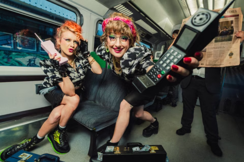 Two women in bold outfits and bright make-up pose energetically on a train, each holding large retro mobile phones. One man in the background reads a newspaper. There is a briefcase with money on the seat.