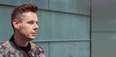 A man with short brown hair and a patterned jacket stands outside in front of a blue-green wall, looking thoughtfully to the left.