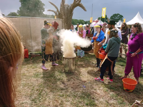 A group of people wearing aprons and protective gear gather around a steaming metal container outdoors at a festival, watching a large cloud of white vapour rise up. Tents and colourful flags are visible in the background.