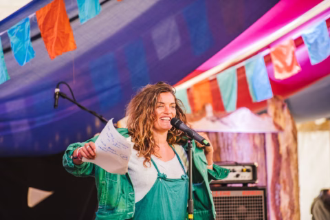 A woman with wavy hair, wearing a green jacket and dungarees, stands smiling at a microphone, holding a sheet of paper. Colourful fabric flags hang above her on a stage with musical equipment in the background.