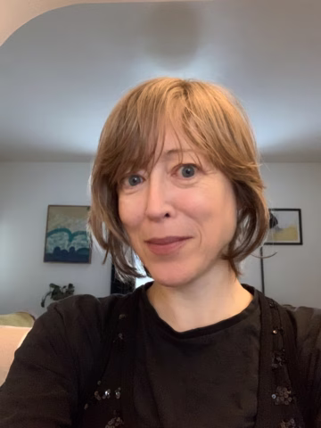 A woman with short, light brown hair and blue eyes smiles slightly at the camera. She is indoors, wearing a black top, with framed art on the wall and a plant in the background.