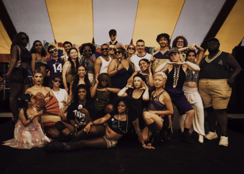 A diverse group of people in colourful, expressive outfits pose together under a large yellow marquee, showing various playful and confident expressions and gestures. The setting suggests a festive or celebratory event.