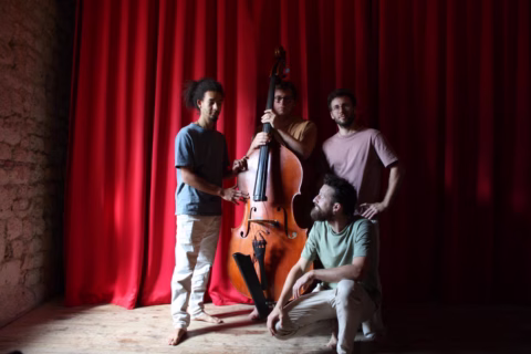 Four men pose together on a wooden stage in front of red curtains. One stands holding a double bass, two others stand beside him, and the fourth is crouched down, looking up at the group.