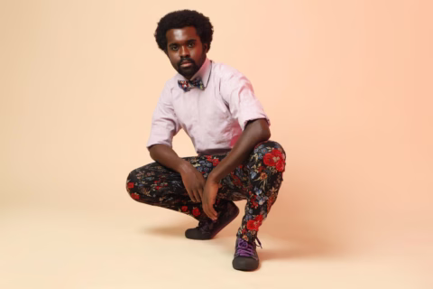 A man with short curly hair squats confidently against a peach background, wearing a light pink shirt, floral bow tie, floral patterned trousers, and black trainers with purple laces.