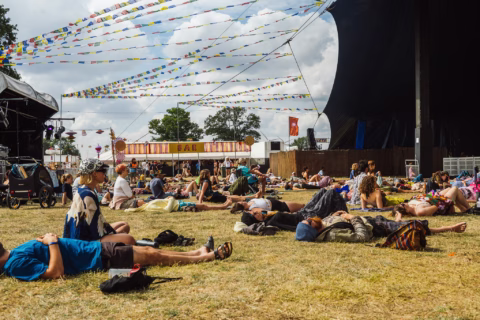 People are lying and relaxing on the grass at an outdoor festival. Colourful bunting is strung overhead, and more people are seen near a bar in the background under a partly cloudy sky.