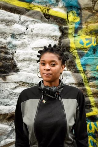A person with short, styled hair and hoop earrings stands in front of a graffiti-covered brick wall, wearing a black and grey jacket and headphones round their neck.