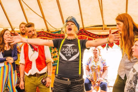 A person in a police-style hat and a “No Justice Just Us” shirt stands with arms outstretched, passionately speaking to a group of people inside a colourful tent. Others watch intently, some smiling or reacting.