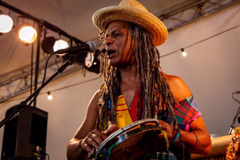 A musician with long dreadlocks and a straw hat sings into a microphone whilst playing a tambourine on stage, illuminated by warm stage lights.