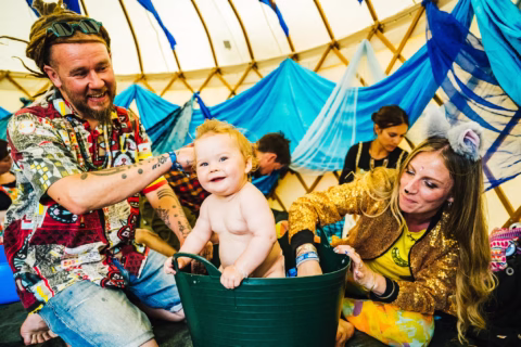 A smiling baby sits in a green tub while a man and a woman, both cheerfully dressed, play with the baby inside a colourful tent decorated with blue fabric. Other adults sit in the background.