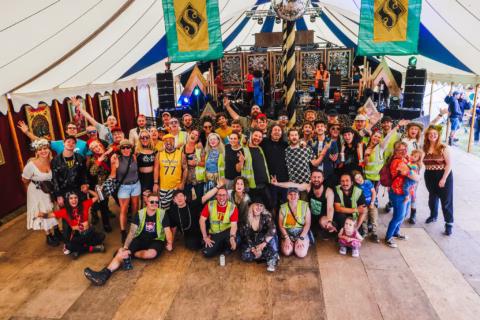 A large, joyful group poses inside a decorated festival marquee, some wearing colourful outfits and safety vests. The background features a stage with banners, lights, and a disco ball overhead.