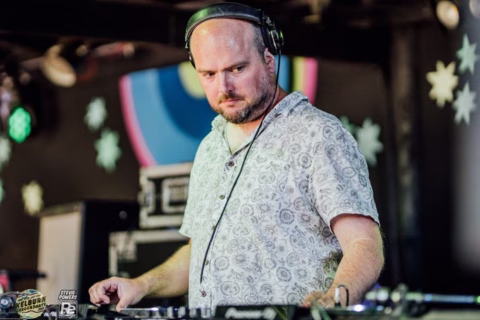 A man wearing headphones and a patterned short-sleeve shirt stands at a DJ booth, focused on mixing music. Colourful, abstract decorations and white star shapes are visible in the background.