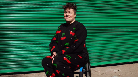 A person with short curly hair wearing a black outfit with red cherries sits in a wheelchair in front of a closed green metal shutter, illuminated by sunlight.