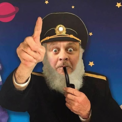 A man in a captain’s costume with a white beard and hat holds a pipe in his mouth and points upwards with a surprised expression. A starry space background with a planet is behind him.
