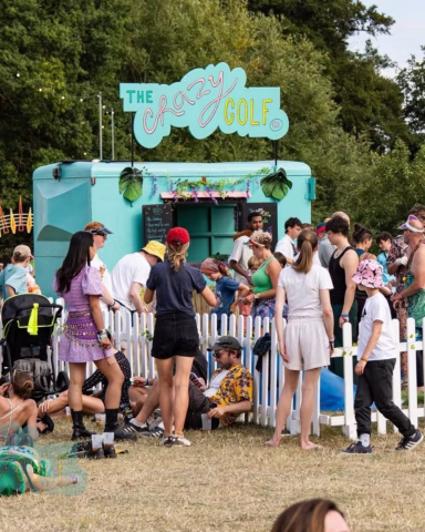 A crowd of people, some standing and some sitting, gathers in front of a teal food van labelled The Crazy Golf at an outdoor event with trees in the background and a white picket fence surrounding the area.