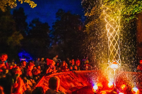 A crowd gathers at night around a brightly lit water fountain with swirling streams and glowing red-orange lights, surrounded by trees and festive atmosphere.