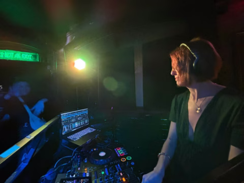 A DJ wearing headphones stands at a booth with mixing equipment and a laptop, looking towards a dimly lit dance floor where a green neon sign glows and people are faintly visible.