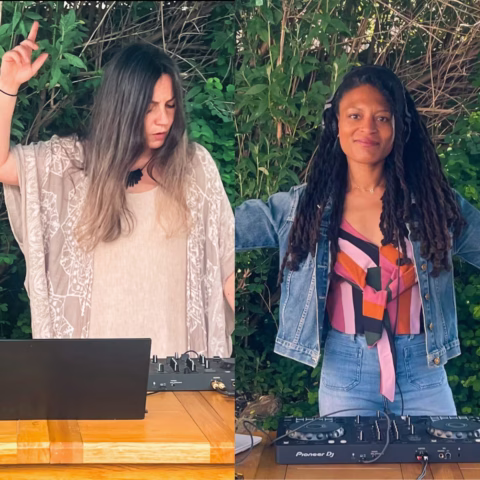 Two women DJ outdoors surrounded by green foliage. One wears a flowy beige top with her eyes closed and hand raised, while the other, in headphones and a denim jacket, smiles at the camera. Both stand behind DJ equipment.