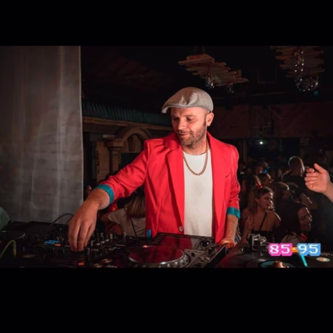 A DJ in a red jacket and white cap mixes music at a lively indoor party, with people dancing in the background and neon 85-95 text in the lower right corner.