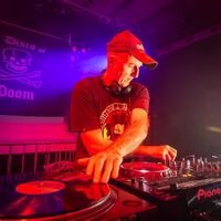 A DJ wearing a red cap and headphones is mixing music on turntables and a mixer, illuminated by colourful stage lights, with a skull and crossbones banner in the background.