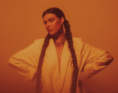 A woman with long plaited hair, wearing a white blazer and a statement necklace, stands confidently with her hands on her hips in warm, orange lighting.