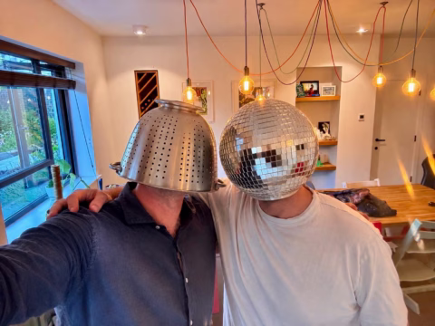Two people stand indoors with their arms round each other. One wears a colander on their head, the other wears a disco ball. Colourful wires hang from the ceiling, and a dining area is visible in the background.
