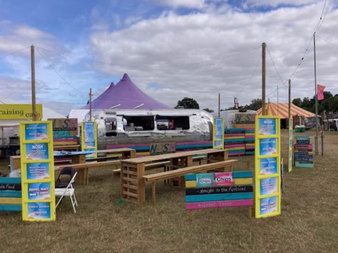 Outdoor food stall with a shiny trailer, wooden benches, colourful signs, and a purple tent in the background, set up on grass under a partly cloudy sky at a festival.