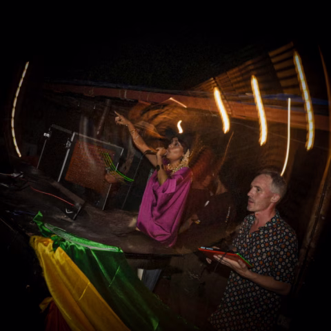 A woman in a purple outfit sings passionately into a microphone whilst another person in a patterned shirt stands nearby under colourful lights and a draped flag at a lively nighttime event.