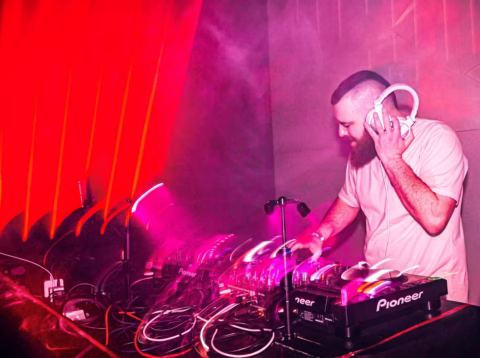 A DJ with a beard and headphones stands behind a mixing desk in a club, surrounded by pink and red lighting, adjusting controls as he performs.