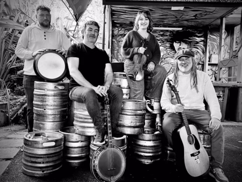 Four people sit and stand on beer kegs, holding musical instruments—a banjo, violin, drum, and guitar—smiling for the camera in an outdoor setting with plants and art in the background.