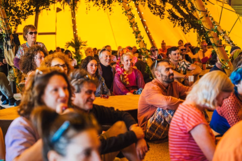 A group of people sit closely together inside a yellow tent, attentively watching something out of view. The tent is decorated with lights and greenery, creating a warm, festive atmosphere.