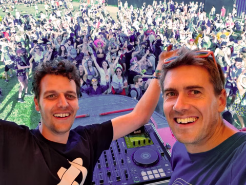 Two smiling men take a selfie on stage in front of DJ equipment, with a large, cheering crowd enjoying an outdoor music festival in the background on a sunny day.