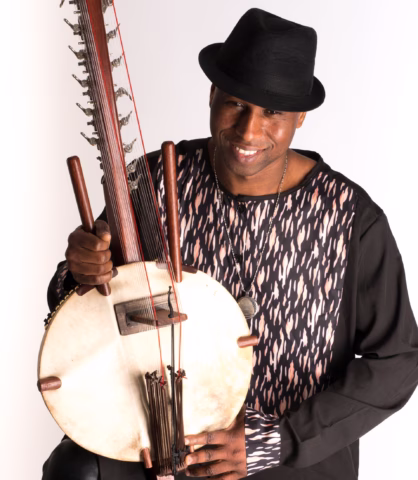 A smiling man in a black hat and patterned shirt holds a traditional African kora instrument, posing against a plain white background.