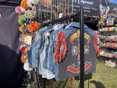 A rail of denim waistcoats is displayed outdoors, with one featuring a large Guns N’ Roses Appetite for Destruction patch. Colourful artificial flowers and festival merchandise are visible in the background.