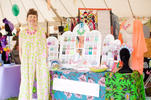 A smiling woman in a bright floral dress stands at a colourful jewellery stall, displaying handmade earrings and necklaces, inside a lively outdoor market marquee.