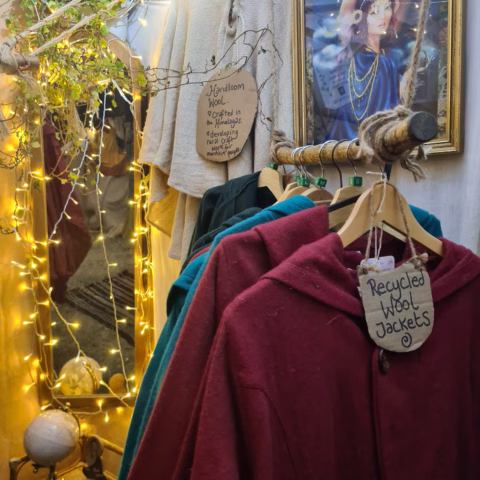 A clothing rail with colourful wool jackets, a Recycled Wool Jackets sign, fairy lights, a potted plant, and a framed portrait in the background at a cosy indoor market stall.